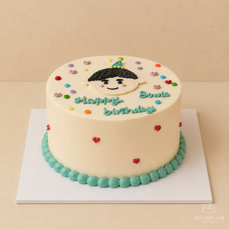 Front view of a little boy birthday cake with hand-piped face, blue cream border and red heart details