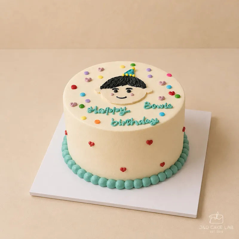 Front view of a little boy birthday cake with hand-piped face, blue cream border and red heart details