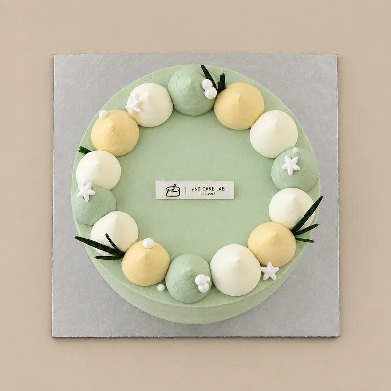 Top view of Lychee Green Tea Cloud cake with a ring of ivory, pale yellow and green cream dollops and small white flowers.