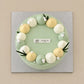 Top view of Lychee Green Tea Cloud cake with a ring of ivory, pale yellow and green cream dollops and small white flowers.