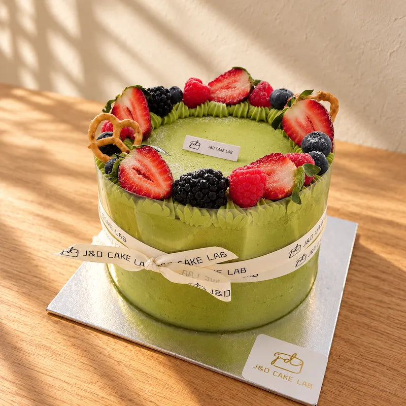 Angled view of J&D Cake Lab’s Uji matcha cream cake with strawberries, mixed berries, pretzels and an ivory ribbon.