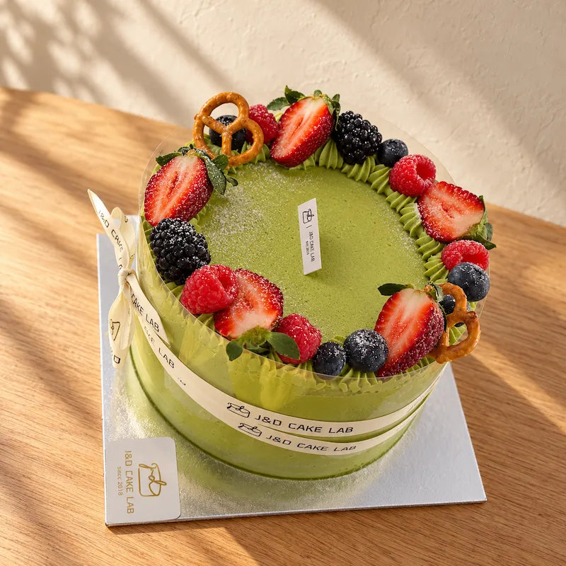 J&D Cake Lab Uji matcha cream cake with green frosting, fruit decorations and a branded ribbon bow on a silver cake board.